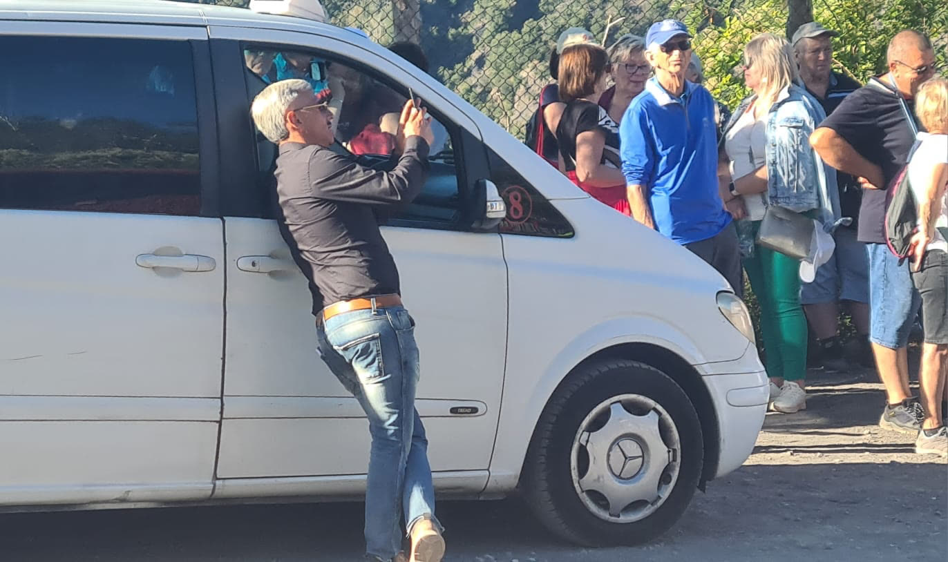 Giovanni leaning against a white minibus, checking his phone while tourists queue behind him